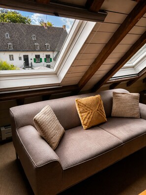 A room with a sloped ceiling, a brown sofa with cushions, and a view of a building through a skylight. - Hotel Winselerhof by Flow (Landgraaf)