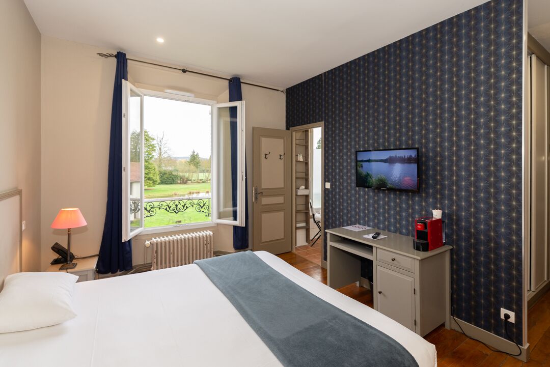 Ein Hotelzimmer mit Bett, Schreibtisch, Fernseher und Blick auf einen Garten durch ein Fenster.