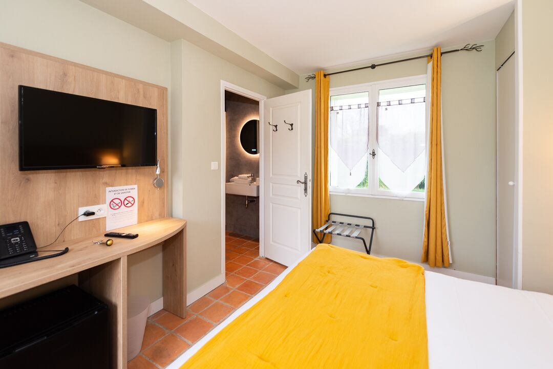 A hotel room with a bed, a desk with a TV, a bathroom, and a window with curtains.