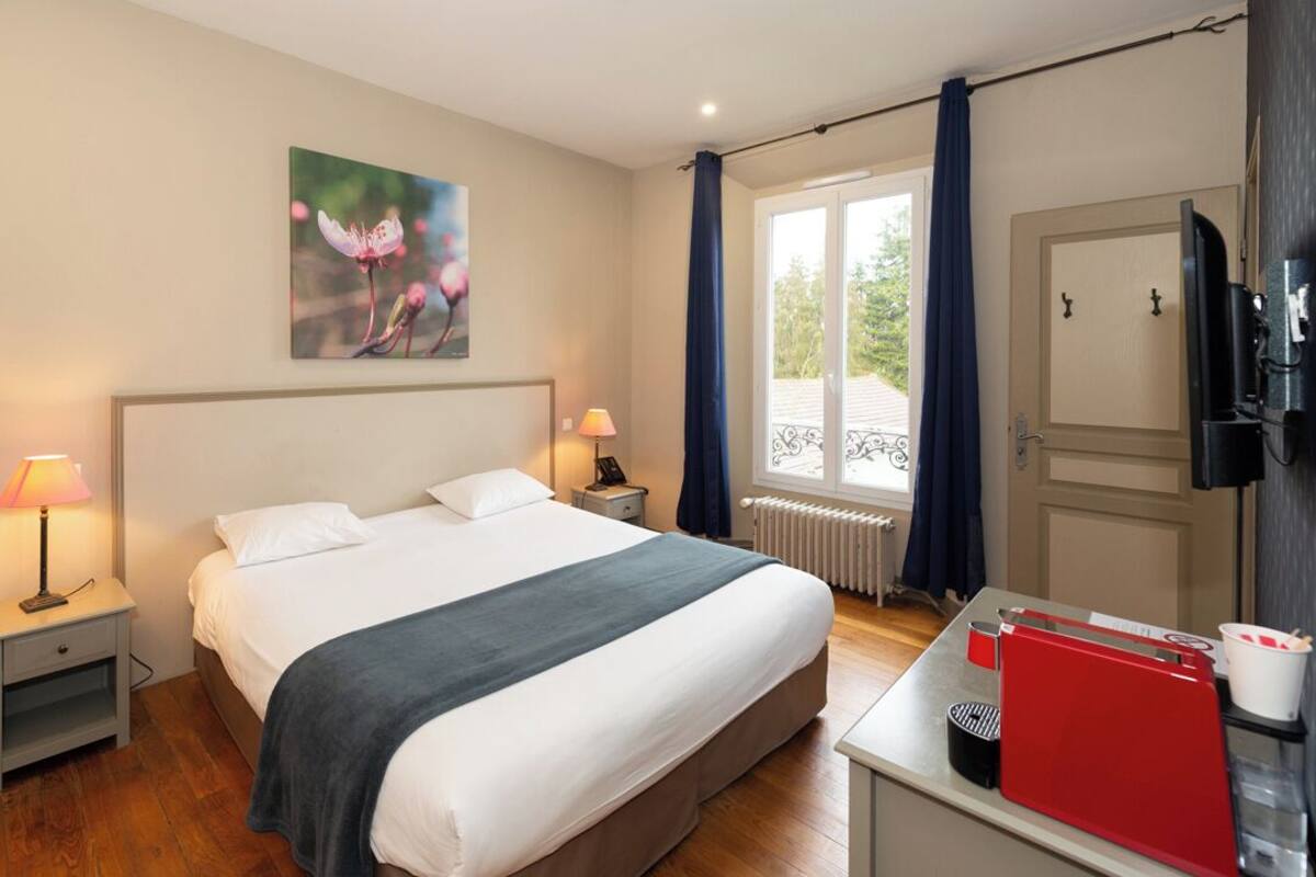 Hotelkamer met een bed, nachtlampjes, een raam met gordijnen, een radiator, een televisie en een bureau met een rode map en een kop.