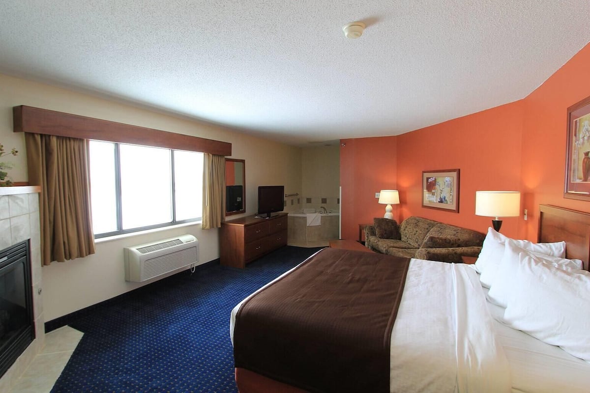 Ein Hotelzimmer mit einem großen Bett, einem Schreibtisch, einem Sessel, einer Couch, einem Kamin, einem Fenster mit Vorhängen und einem durch eine offene Tür einsehbaren Badezimmer.