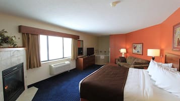 Quarto de hotel com uma cama grande, escrivaninha, cadeira, sofá, lareira, janela com cortinas e banheiro visível por uma porta aberta.