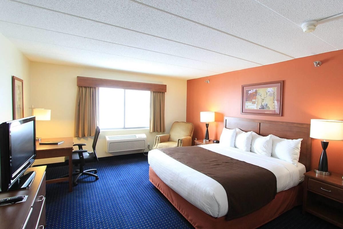 Ein Hotelzimmer mit einem großen Bett, einem Schreibtisch mit Fernseher, einem Sessel und einer Schreibtischlampe.