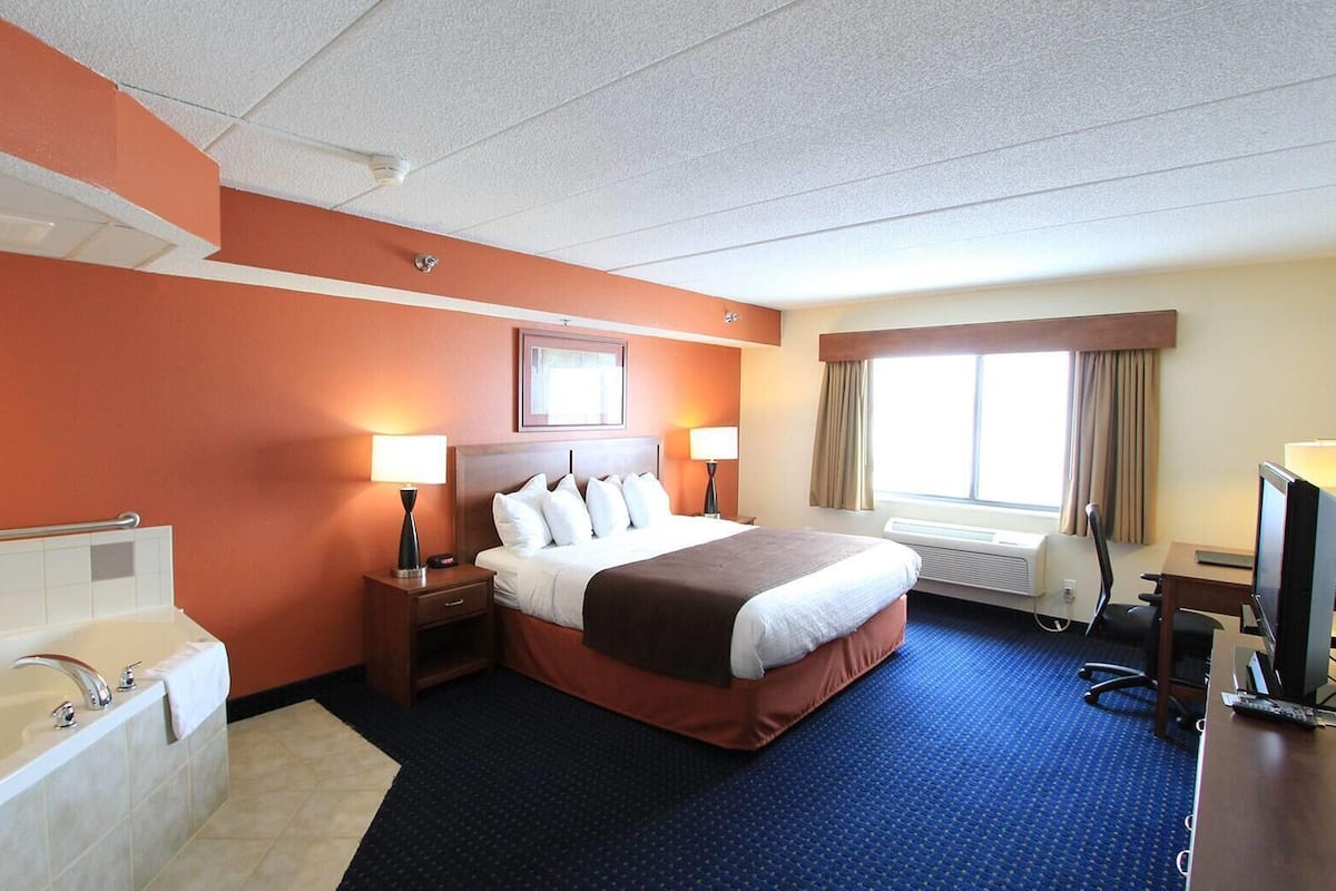 Ein Hotelzimmer mit einem großen Bett, einem eigenen Badezimmer mit Badewanne, einem Schreibtisch mit Computer und einem Fenster mit Vorhängen.