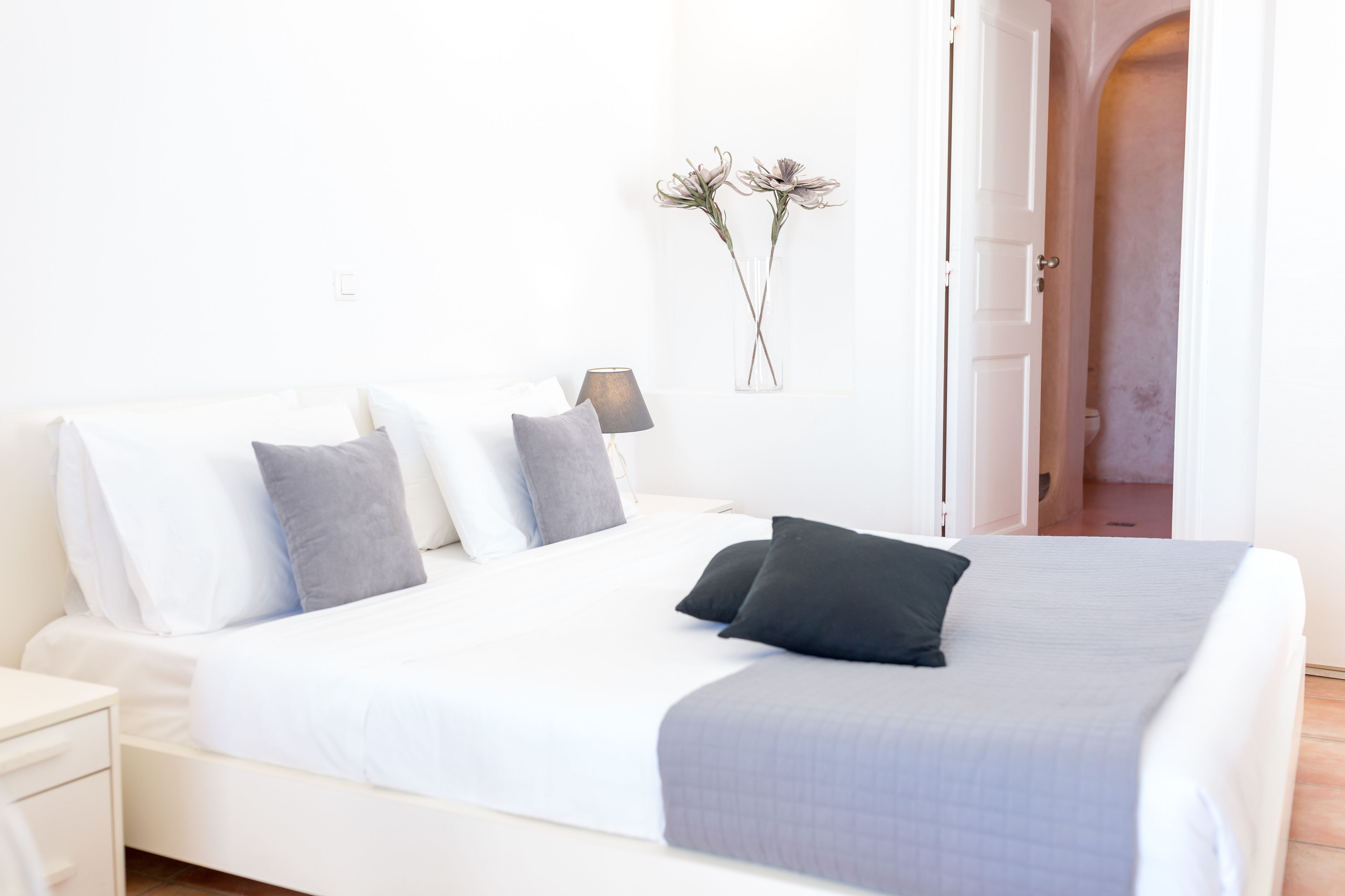 A neatly made bed with a white and gray bedspread, two black pillows, and a vase with dried flowers on the wall.