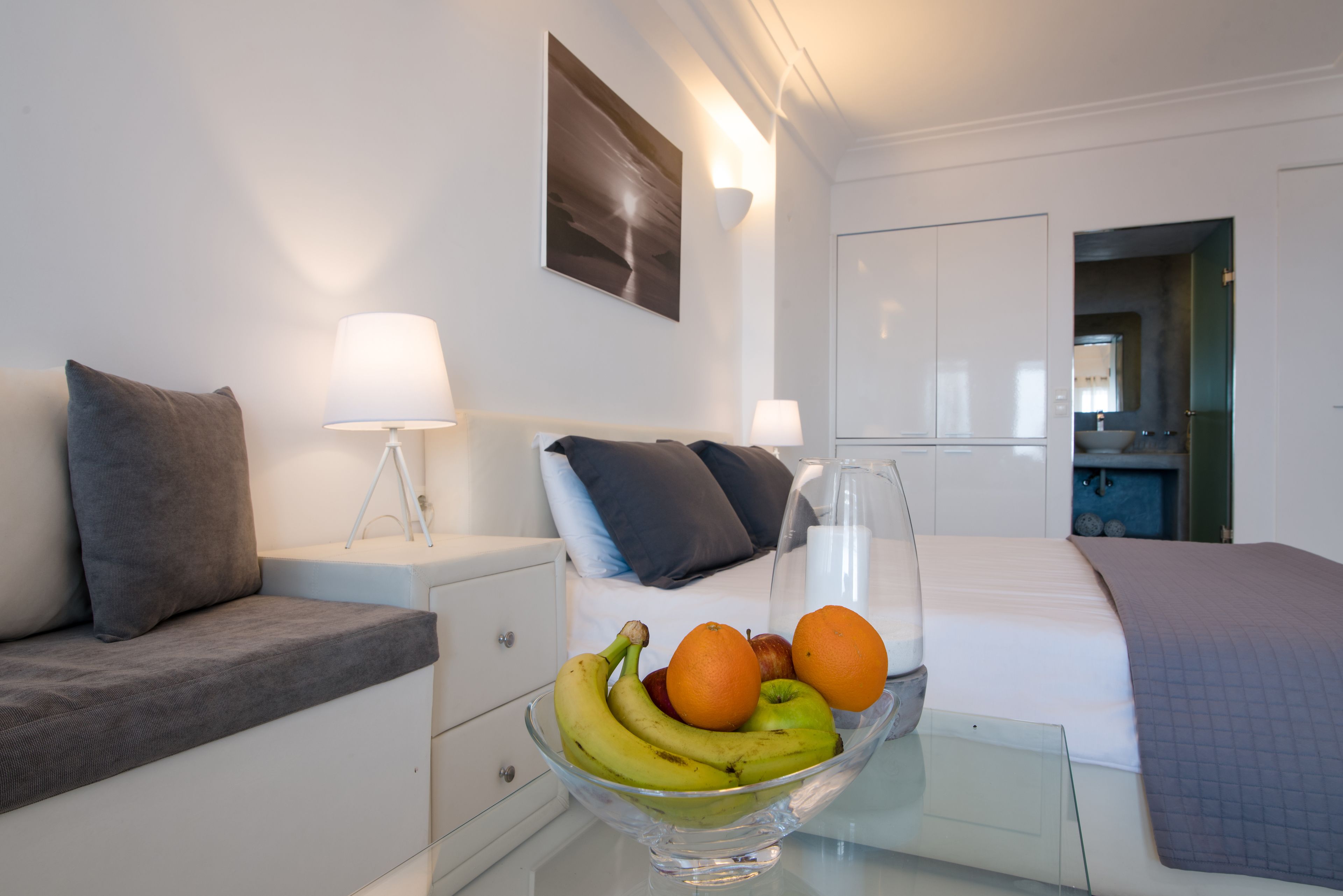 A hotel room with a bed, a bedside table with a lamp, a glass bowl of fruit, and a painting on the wall.