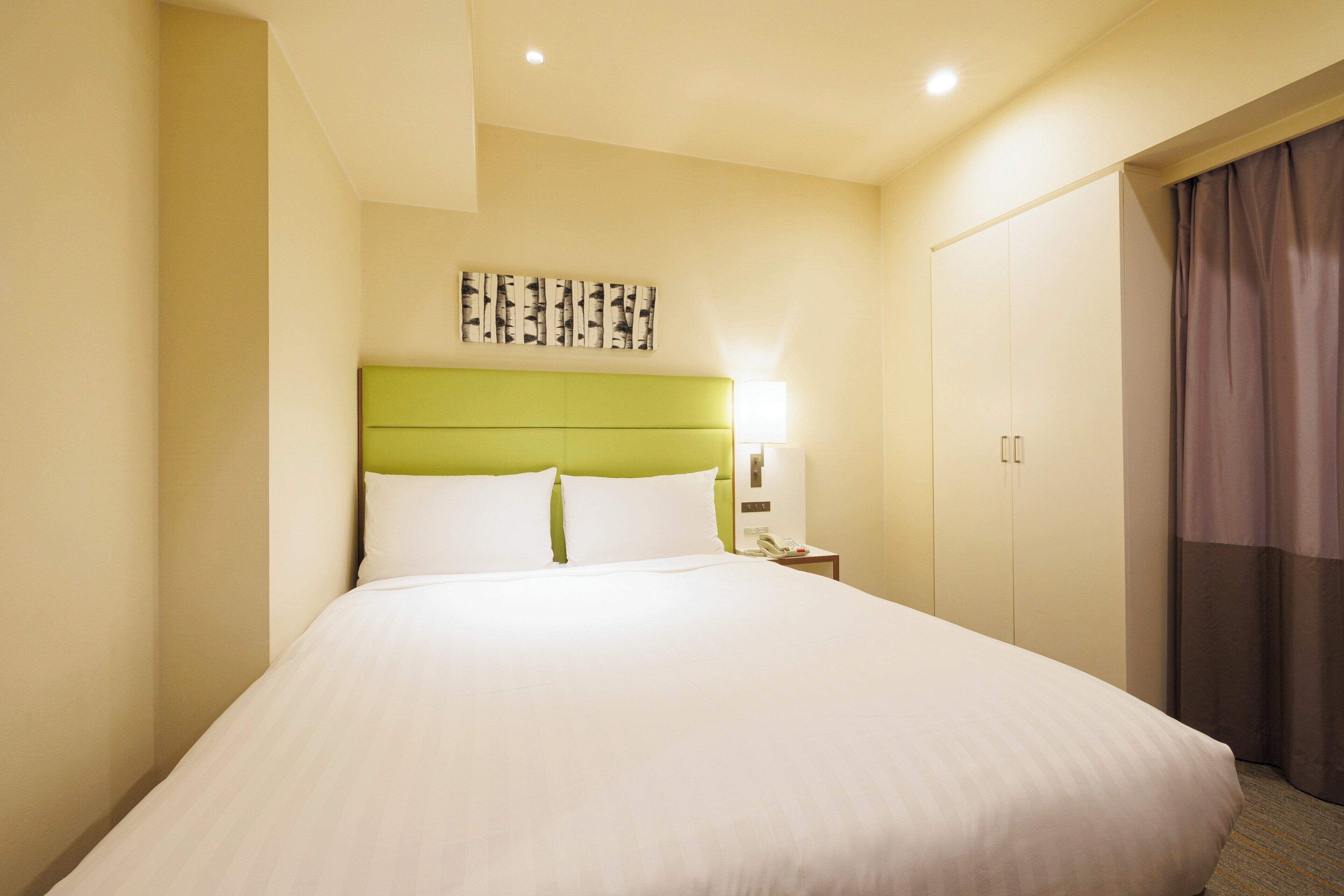 A hotel room with a large bed, a green headboard, a bedside lamp, a telephone, and a wardrobe.
