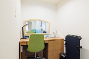 Una camera d'albergo con una scrivania in legno, una sedia verde, uno specchio, un tavolino con una lampada, una sveglia e una valigia.