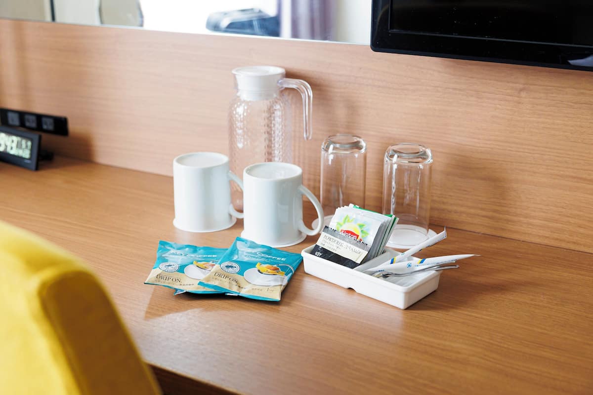 A hotel room desk with a coffee maker, two cups, a glass pitcher, a tray with coffee packets, and a clock.