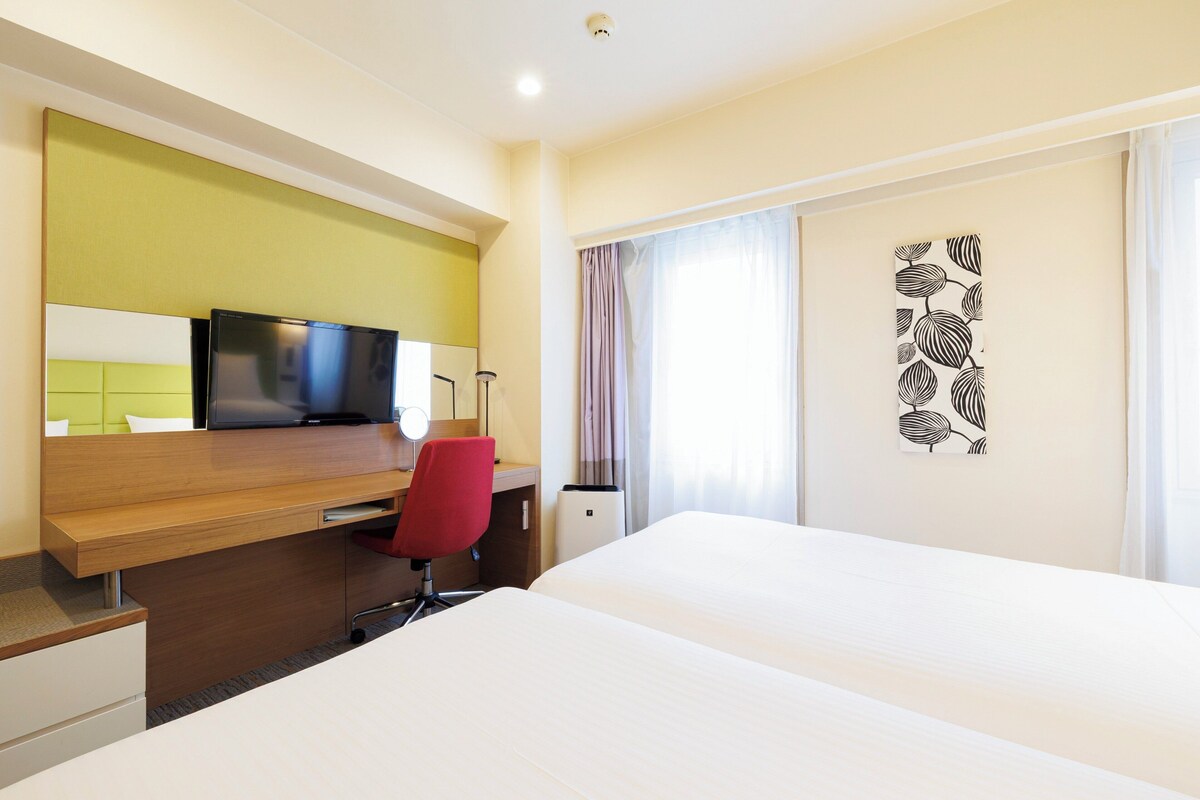 Una camera d'albergo con un letto, una scrivania con un computer, una sedia rossa e una televisione.