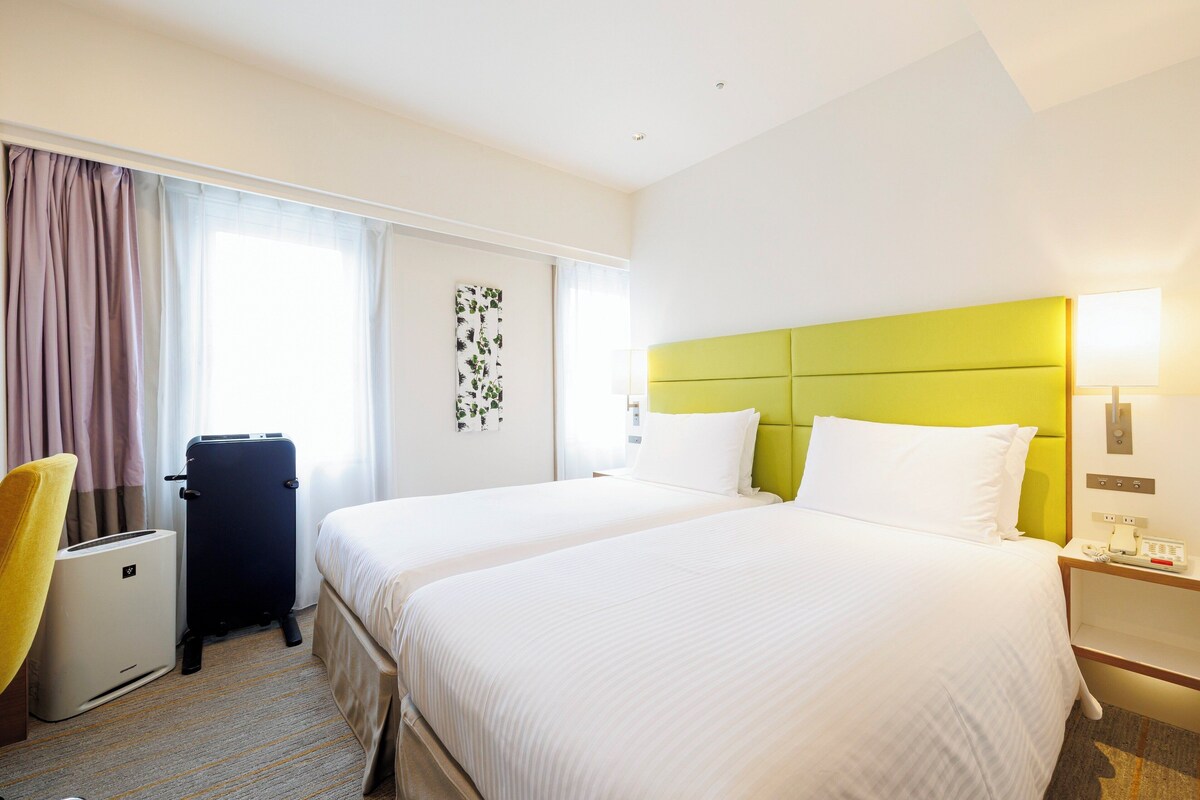 Une chambre d’hôtel avec deux lits, un bureau, une chaise, une valise et une lampe fixée au mur.