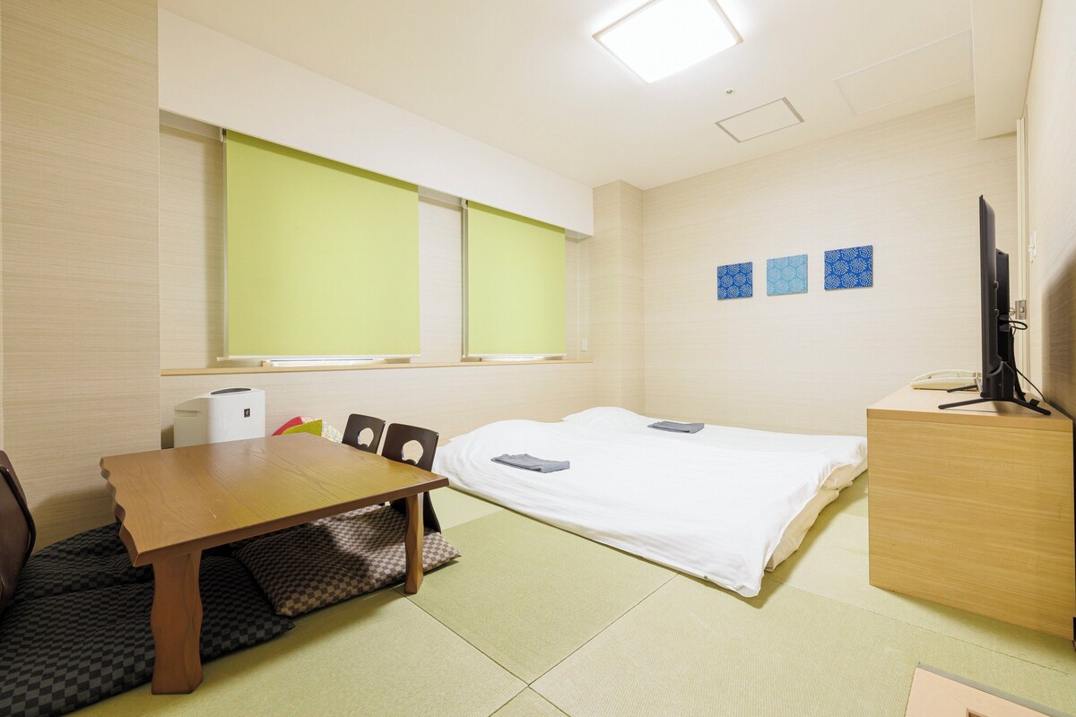 Una camera d'albergo con un letto, una scrivania, una sedia, una televisione e una finestra con tende.