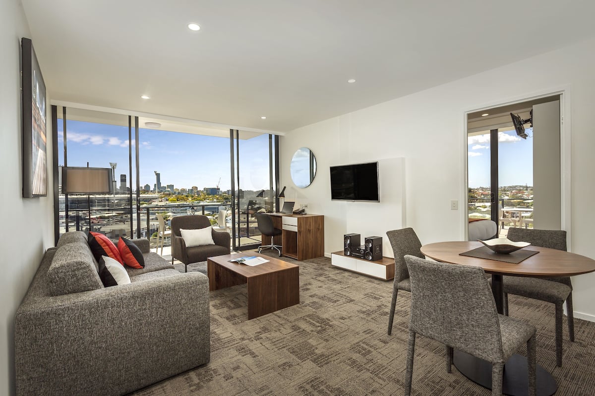 Un salon moderne avec un canapé, une table basse et un coin repas. Un téléviseur est fixé au mur et une grande fenêtre offre une vue sur la ville.