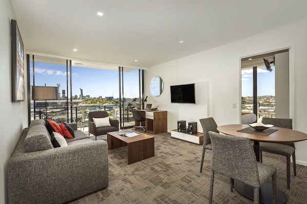 A modern living room with a sofa, coffee table, and dining area. There is a TV mounted on the wall and a large window offering a city view. - Quest Woolloongabba (Woolloongabba)
