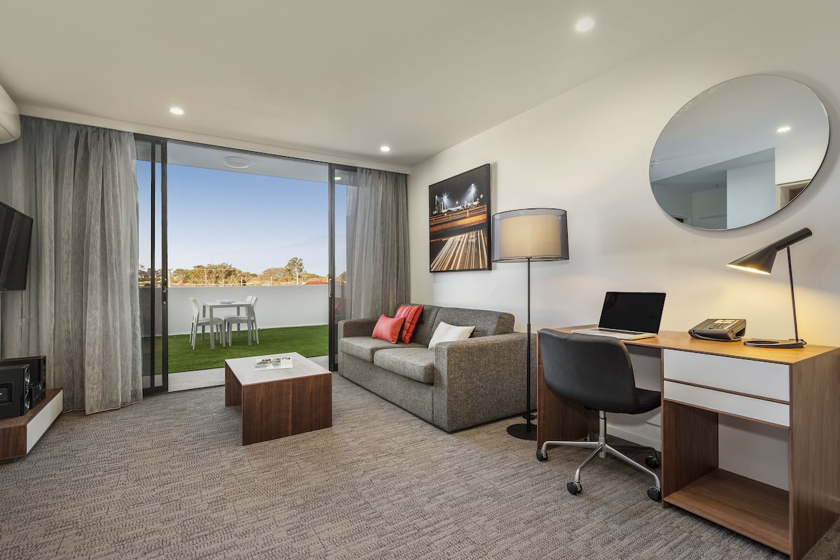 Une chambre d’hôtel moderne équipée d’un canapé, d’un bureau et d’un ordinateur portable, d’un grand miroir et offrant une vue sur le paysage urbain.