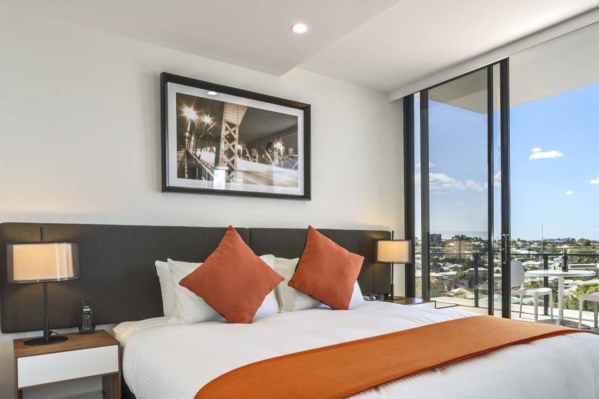Une chambre d’hôtel moderne avec un grand lit, deux tables de chevet avec des lampes, un tableau encadré au mur et une grande fenêtre donnant sur la ville.