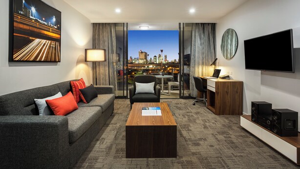 A modern hotel room with a city view, a sofa, a coffee table, a desk, and a TV. - Quest Woolloongabba (Woolloongabba)