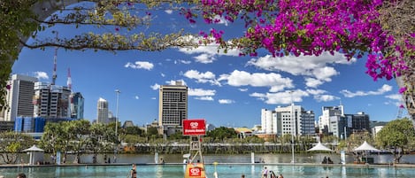 Une piscine située au bord de l’eau, où des personnes se prélassent au soleil et nagent, le tout entouré de bâtiments de la ville et sous un ciel dégagé.