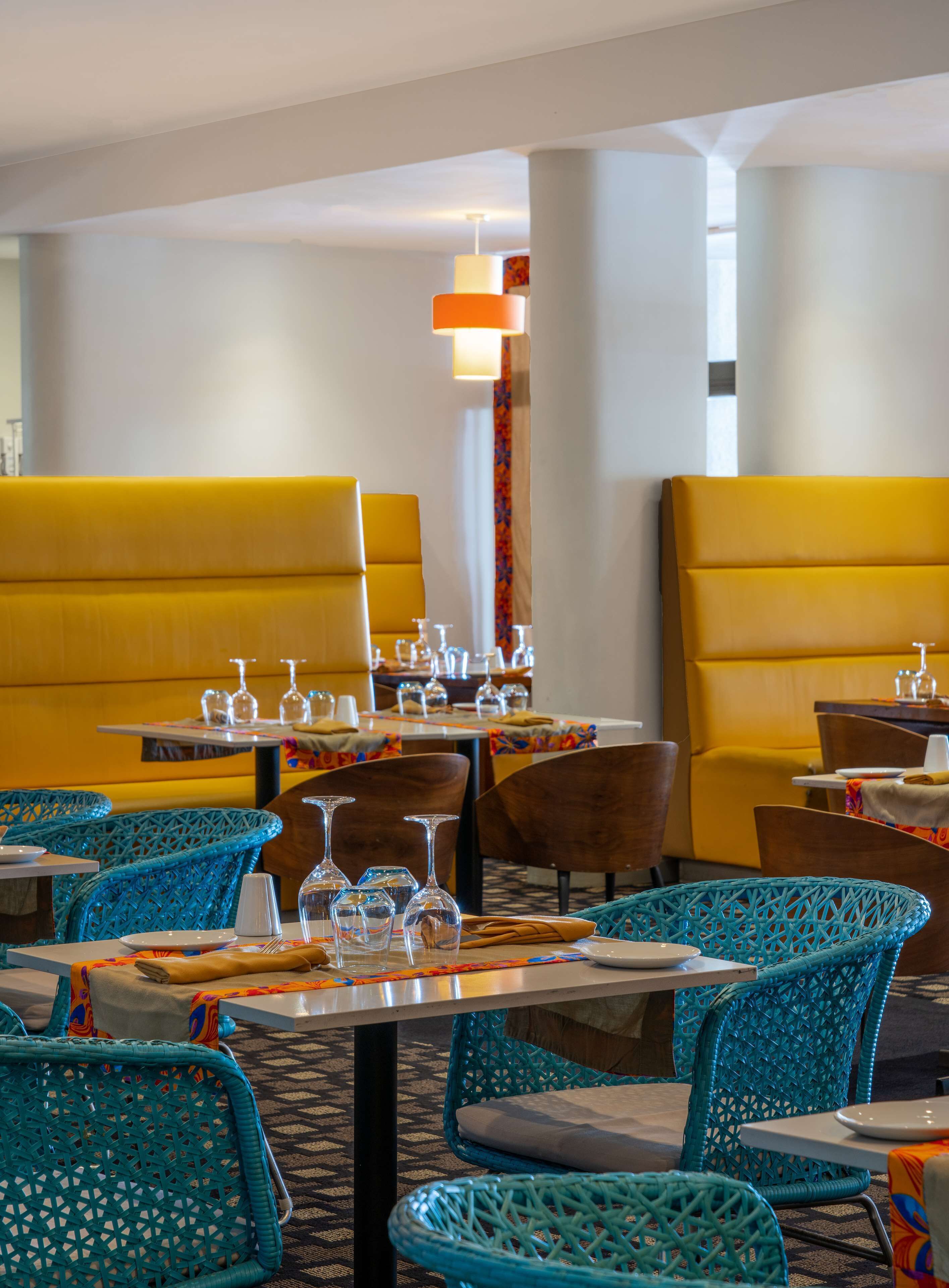 Un restaurant avec des banquettes jaunes et des chaises en osier bleues, des tables dressées avec des verres à vin et des assiettes.