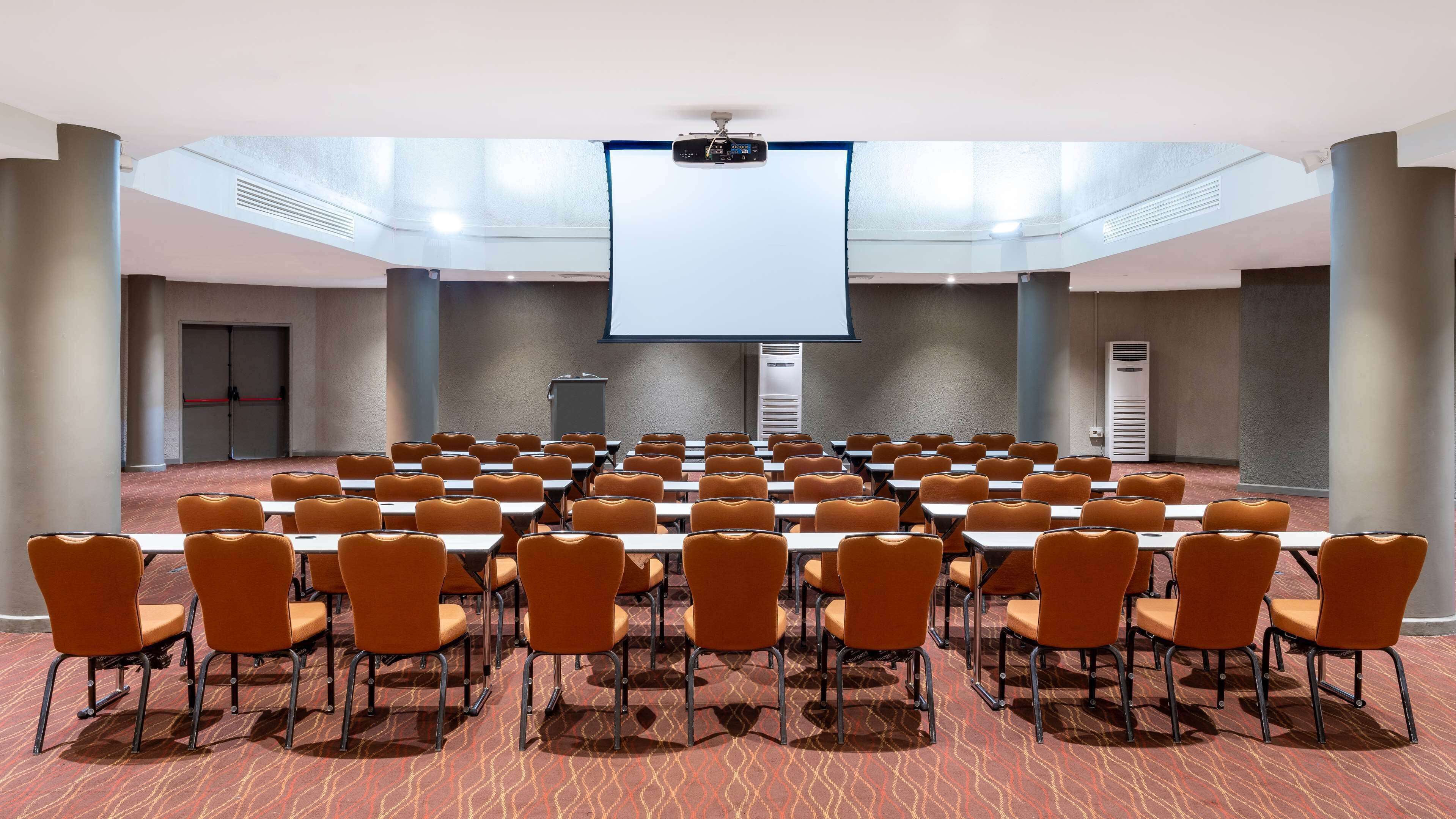 Une salle de conférence avec des rangées de chaises orange et des tables blanches, un grand écran à l’avant et une porte rouge au fond.