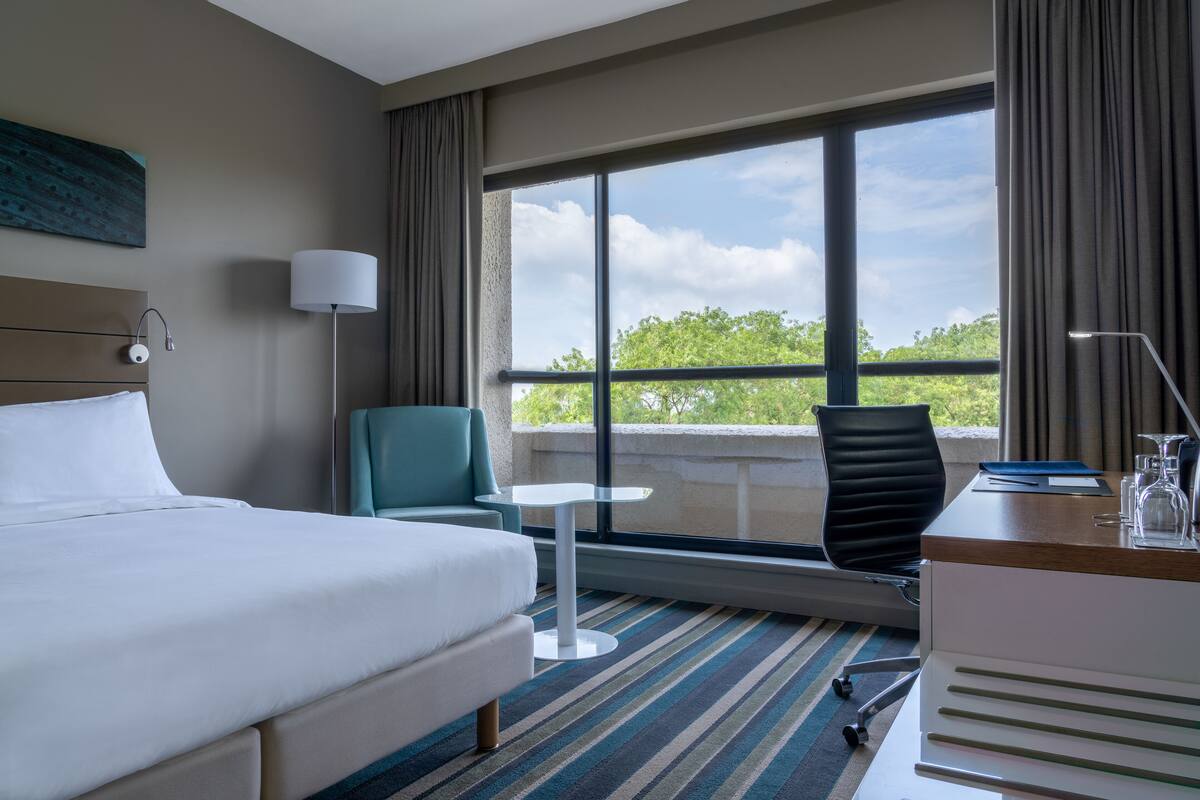 Habitación de hotel con cama, escritorio, silla, mesita pequeña y ventana grande con vista a árboles.