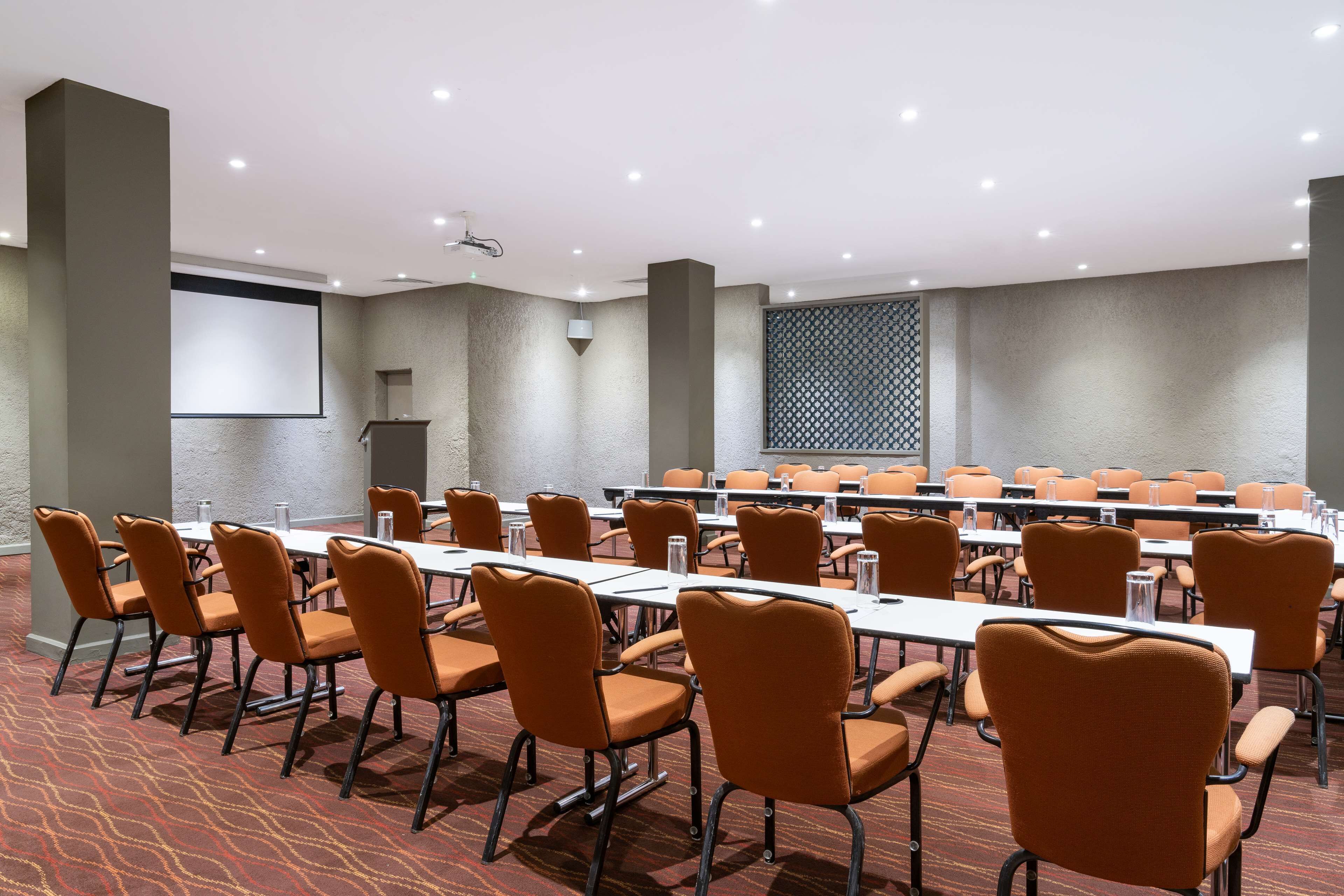 A conference room with rows of orange chairs and white tables, equipped with water bottles and a projector screen.