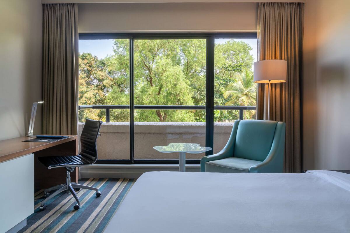 Habitación de hotel con cama, escritorio, silla, una mesita y vistas a árboles a través de un ventanal grande.