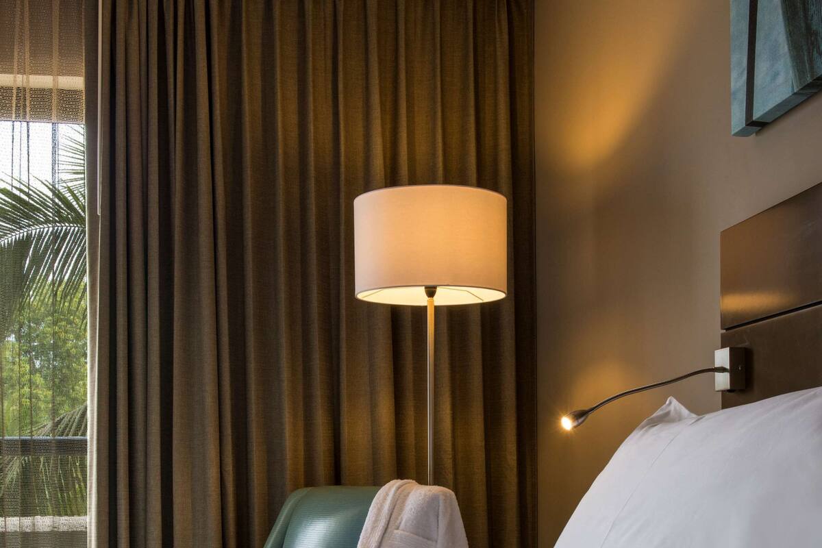 Una habitación de hotel con una cama, un sillón verde, una lámpara y una ventana con cortinas.