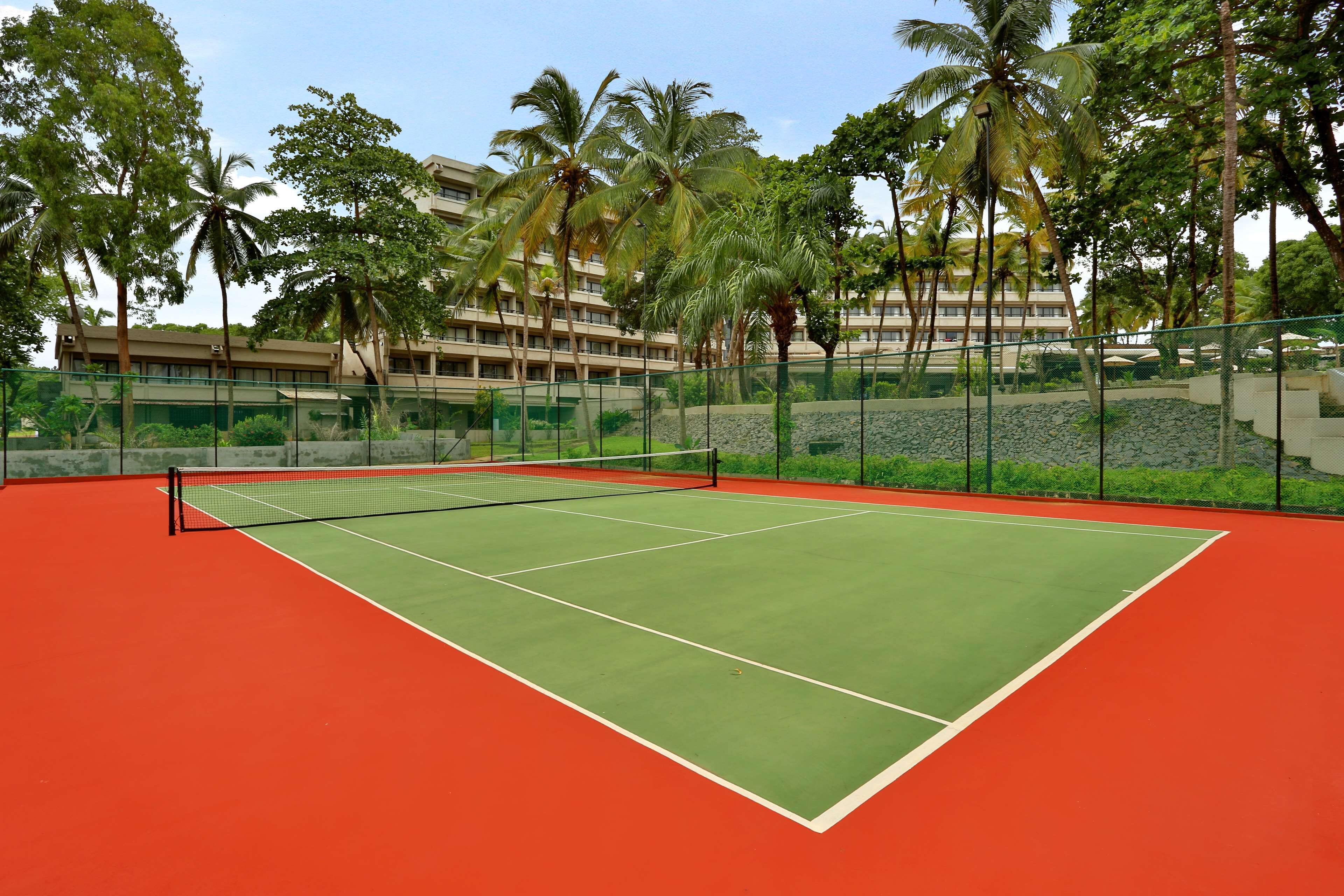 A tennis court with a net, surrounded by a fence, palm trees, and a building in the background.