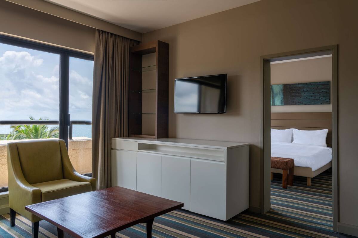 Habitación de hotel con un ventanal grande, un televisor de pantalla plana en la pared, una cama con ropa blanca, un pequeño banco de madera y una mesa.