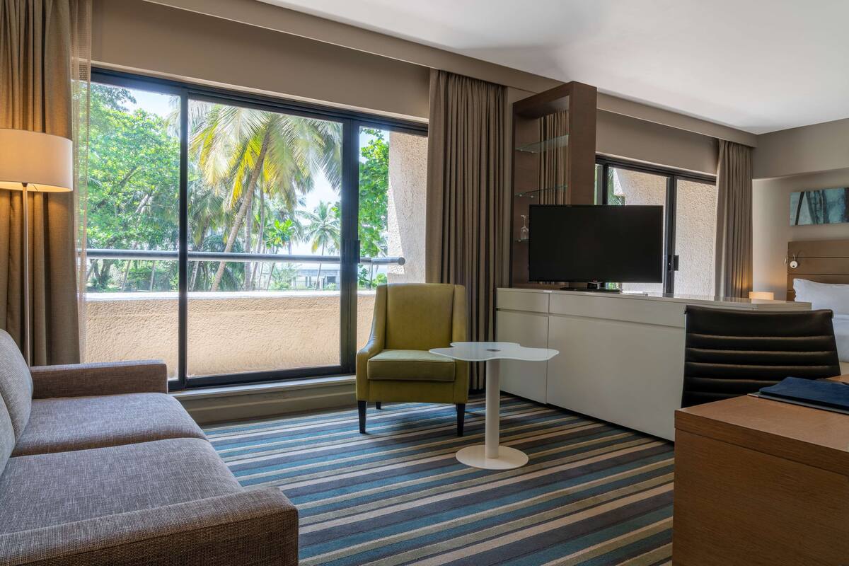 Habitación de hotel con sofá, sillón, mesita, televisor y balcón con vista a palmeras.
