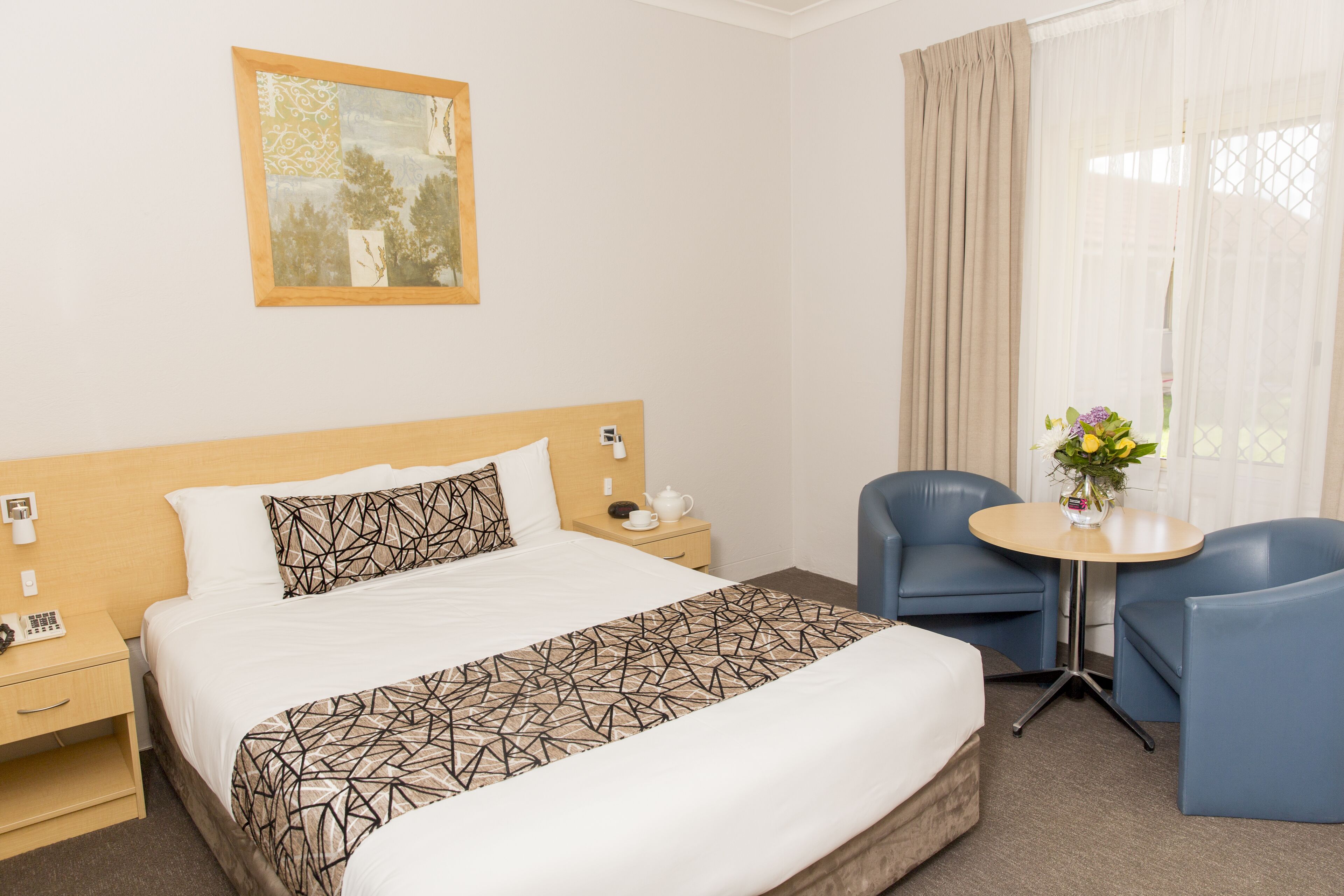 A hotel room with a large bed, a bedside table with a phone, a small table with a vase of flowers, and two blue chairs.