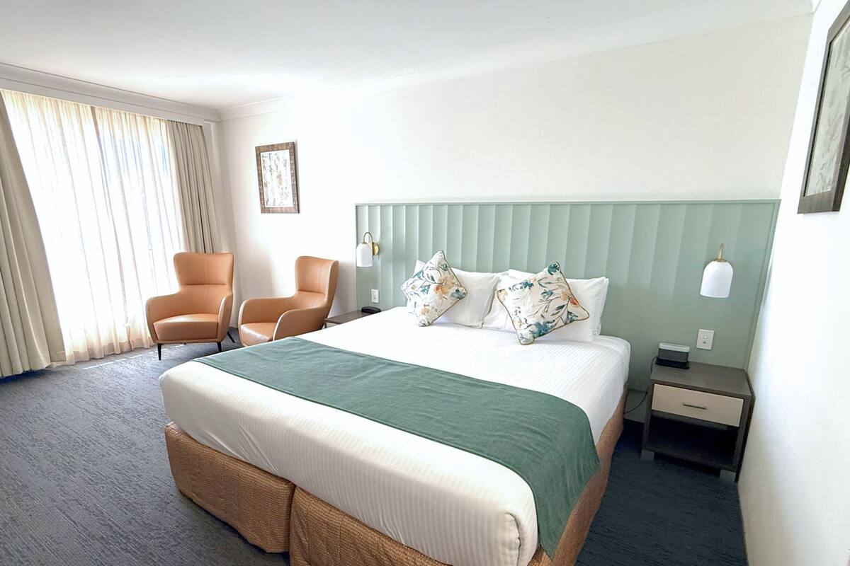 Una habitación de hotel con una cama, dos sillas, una mesita de noche, una lámpara y un ventanal con cortinas.