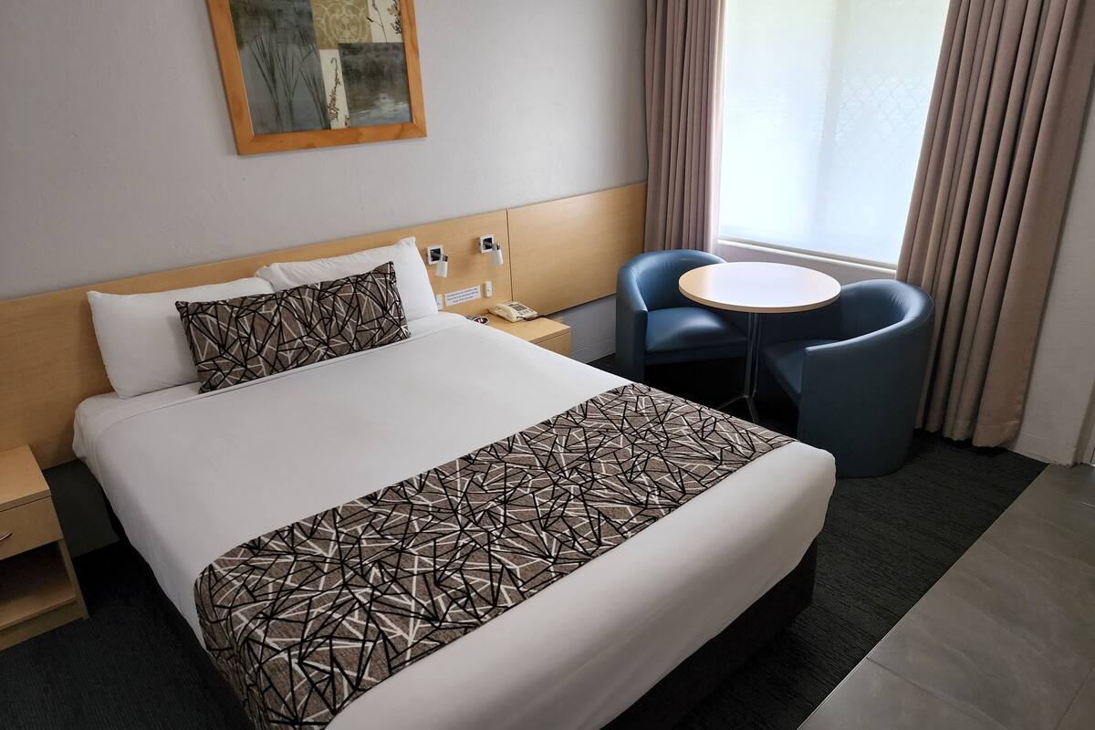 Una habitación de hotel con una cama grande, una mesita de noche con teléfono, una pequeña mesa redonda con una silla y una ventana con cortinas.