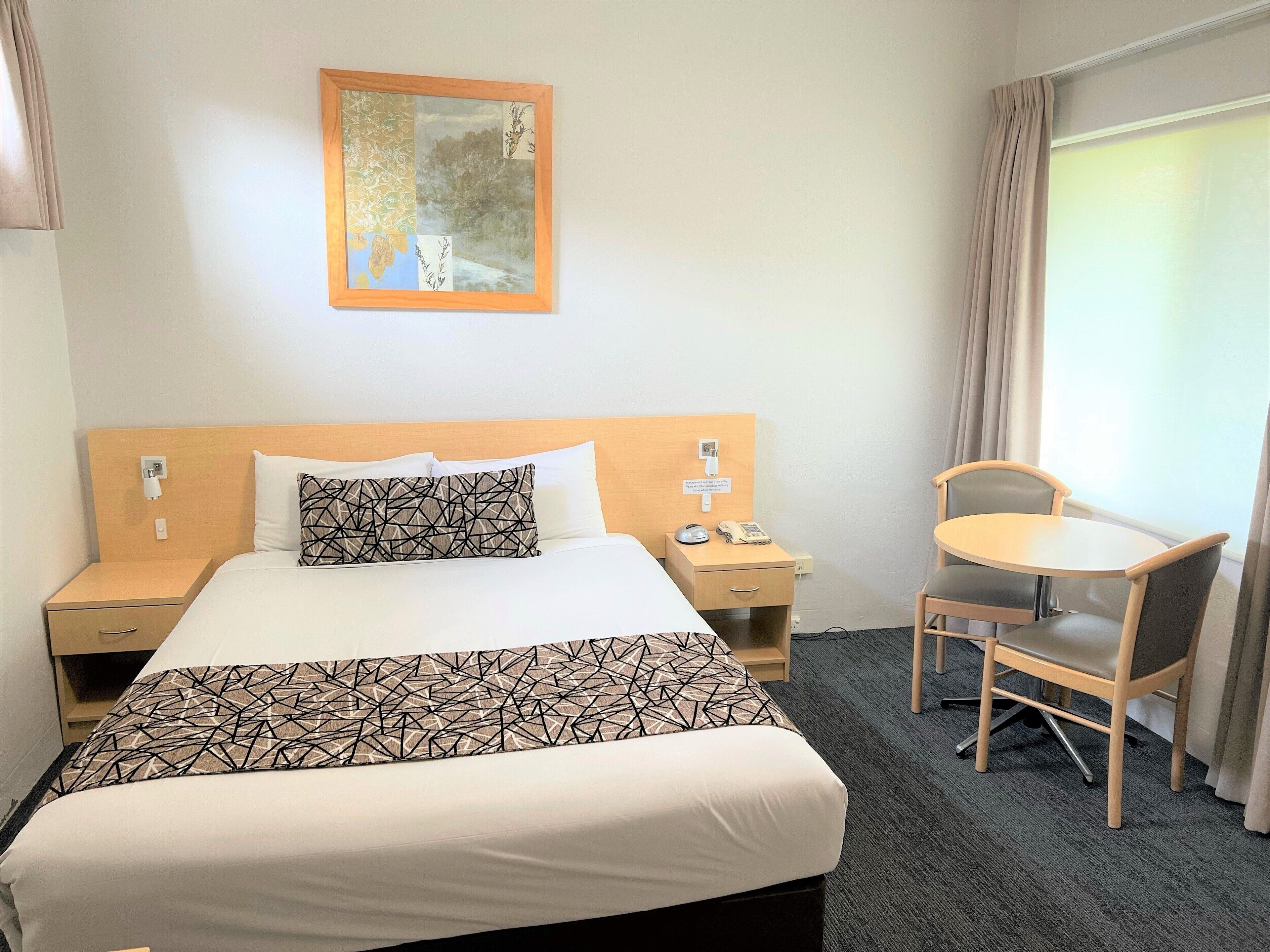A hotel room with a bed, a bedside table with a phone and a lamp, a chair, and a window with curtains.
