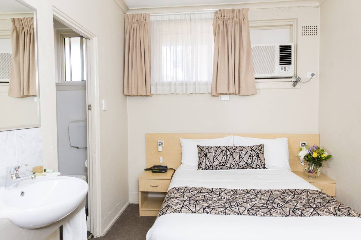 Habitación de hotel con cama, mesita de noche con teléfono y un jarrón de flores, ventana con cortinas y baño con lavamanos y espejo.