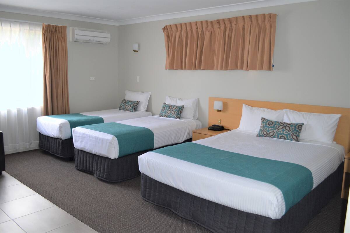 Habitación de hotel con tres camas, cada una con un edredón blanco y azul turquesa, y un ventanal grande con cortinas.