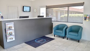 Un espace d’accueil comprenant un comptoir, deux fauteuils bleus, un téléviseur, une horloge et une fenêtre donnant sur un bâtiment et de la verdure.