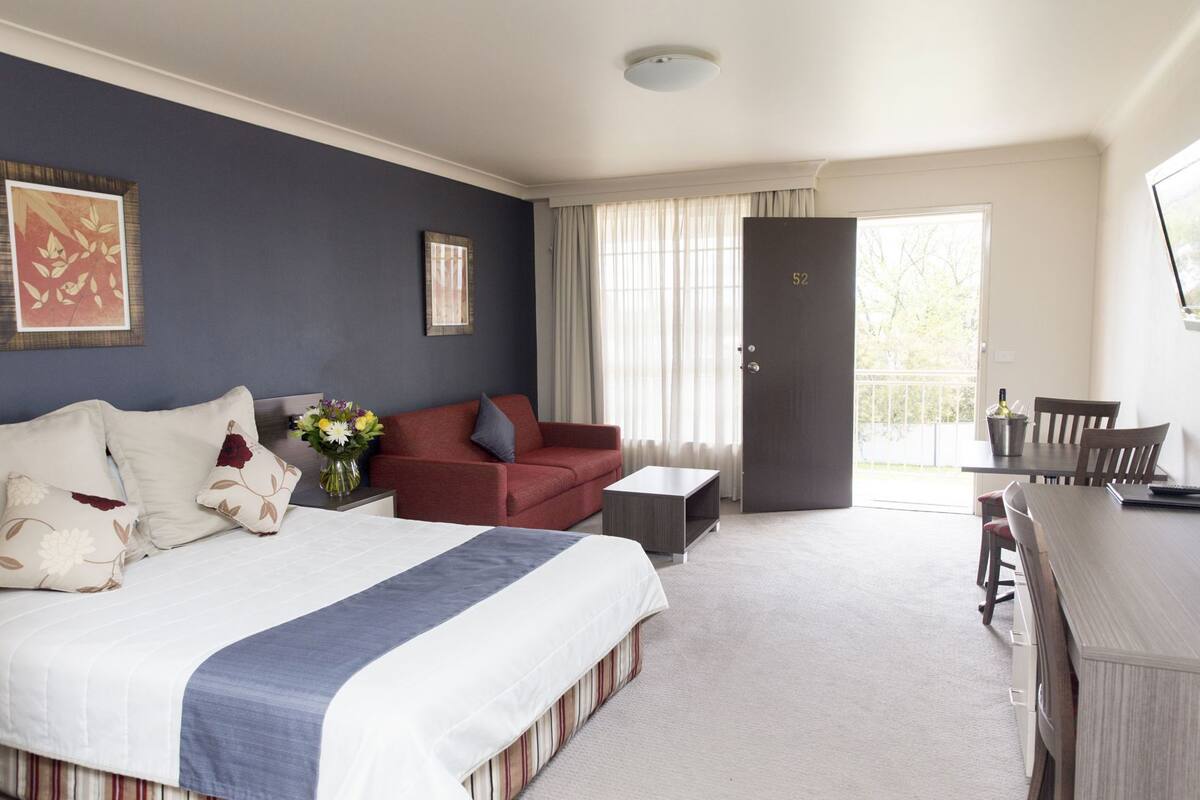 Habitación de hotel con cama, sofá rojo, una mesita y balcón con mesa y sillas.