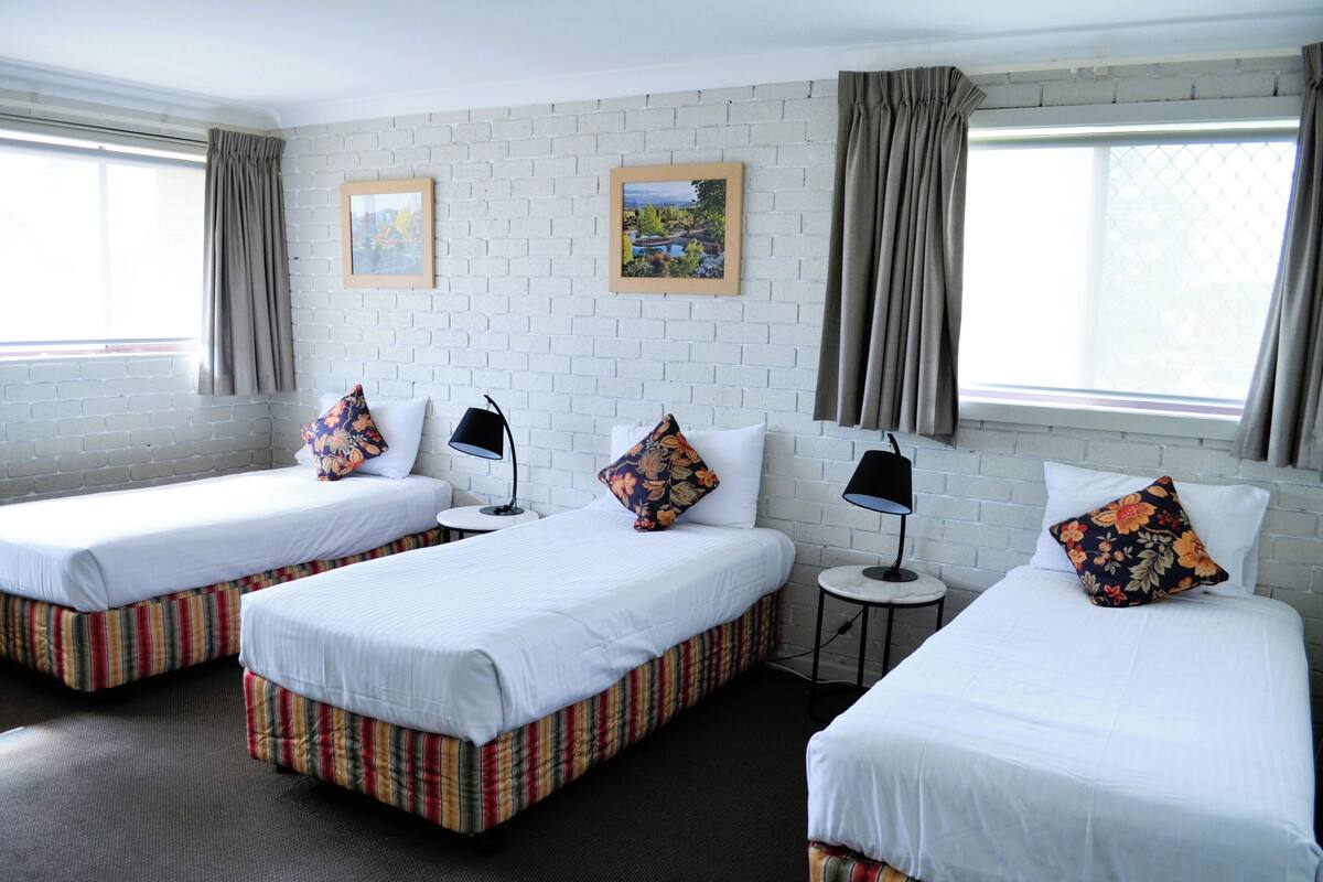 Habitación de hotel con tres camas individuales, cada una con cubrecolchón a rayas, ropa de cama blanca y almohadones decorativos. Hay dos mesitas redondas con lámparas, y las paredes son de ladrillo blanco con dos cuadros enmarcados.