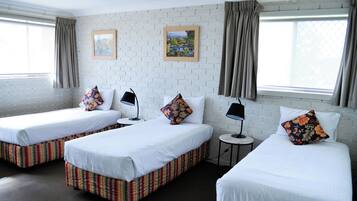 A hotel room with three single beds, each with a striped bed skirt, white linens, and decorative pillows. There are two small round tables with lamps, and the room has white brick walls with two framed pictures.
