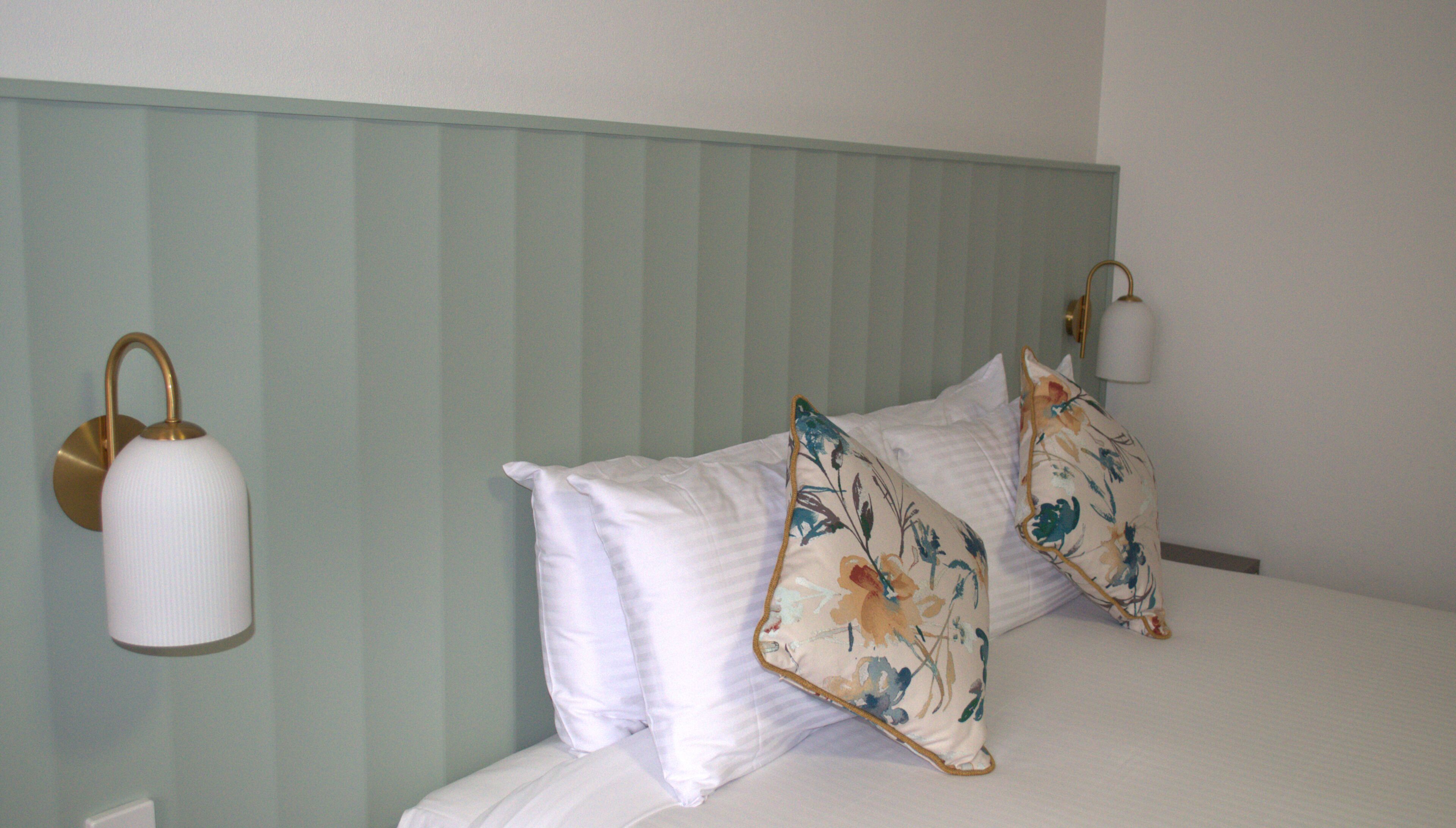 Una cama con ropa de cama blanca y almohadas de estampado floral, una lámpara de pared y un cabecero de color claro.