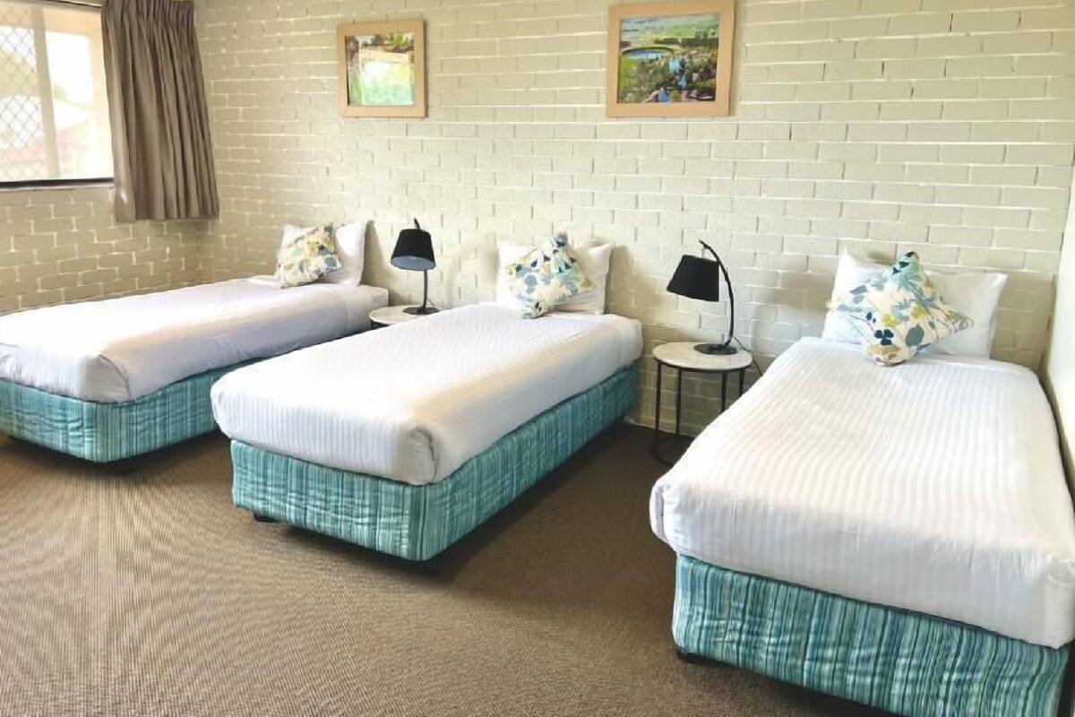 Habitación de hotel con dos camas individuales, una mesita redonda pequeña, una lámpara y una pared de ladrillo con dos cuadros enmarcados.