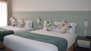 Une chambre d’hôtel avec deux lits, chacun agrémenté d’oreillers à motifs floraux et recouvert d’un plaid vert. On y trouve des lampes fixées au mur et une table de chevet avec un tiroir.