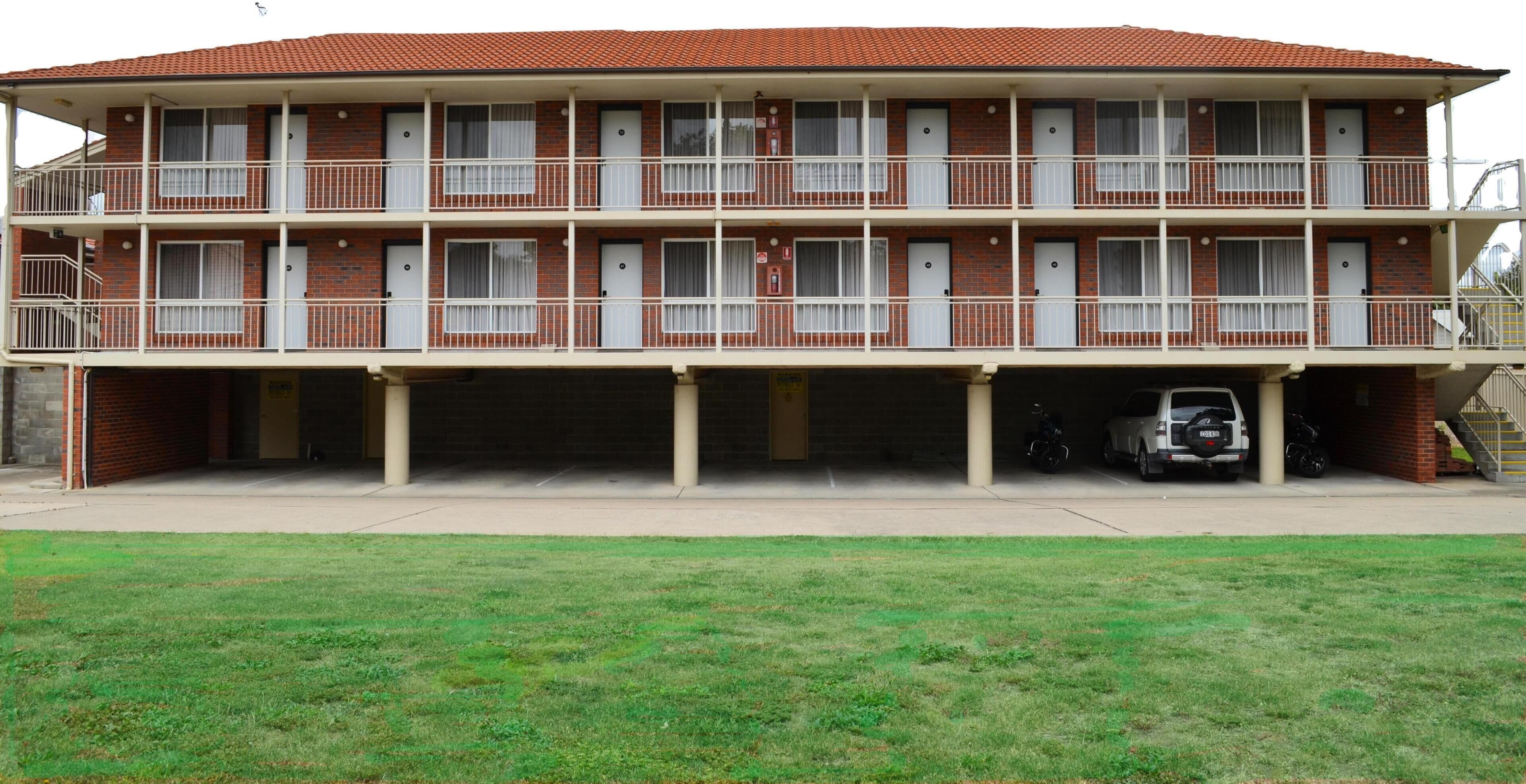 Un edificio de dos pisos con techo plano, varios balcones y un estacionamiento al frente.