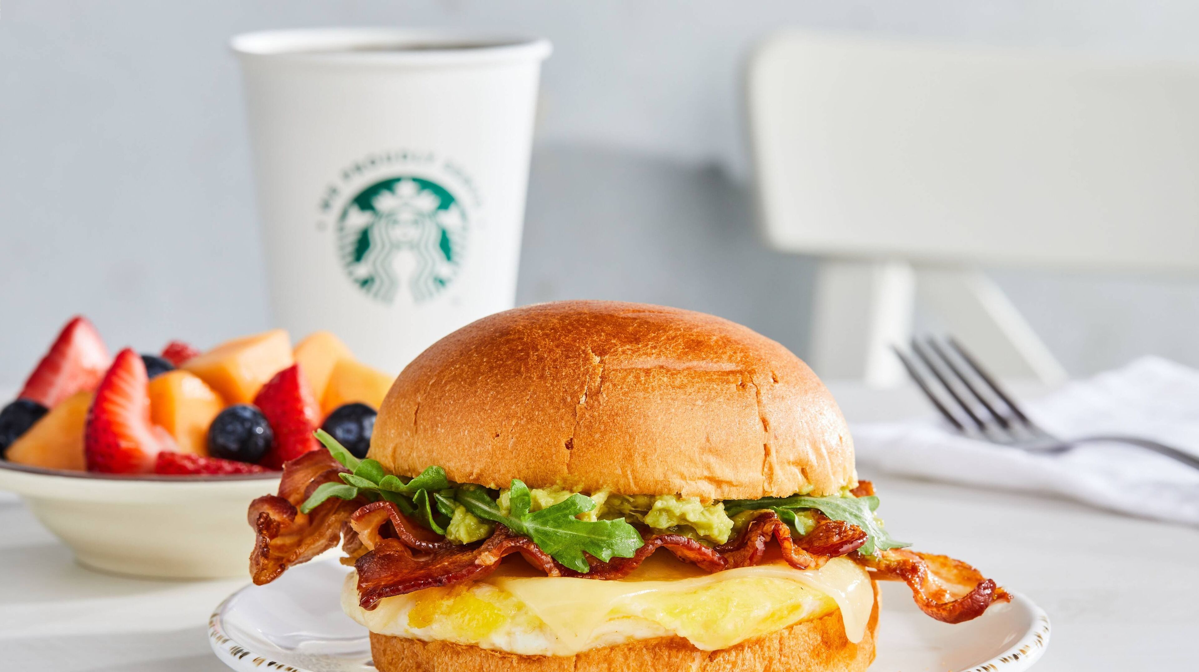 A breakfast sandwich with bacon, lettuce, and cheese on a bun, accompanied by a bowl of fruit and a Starbucks cup.