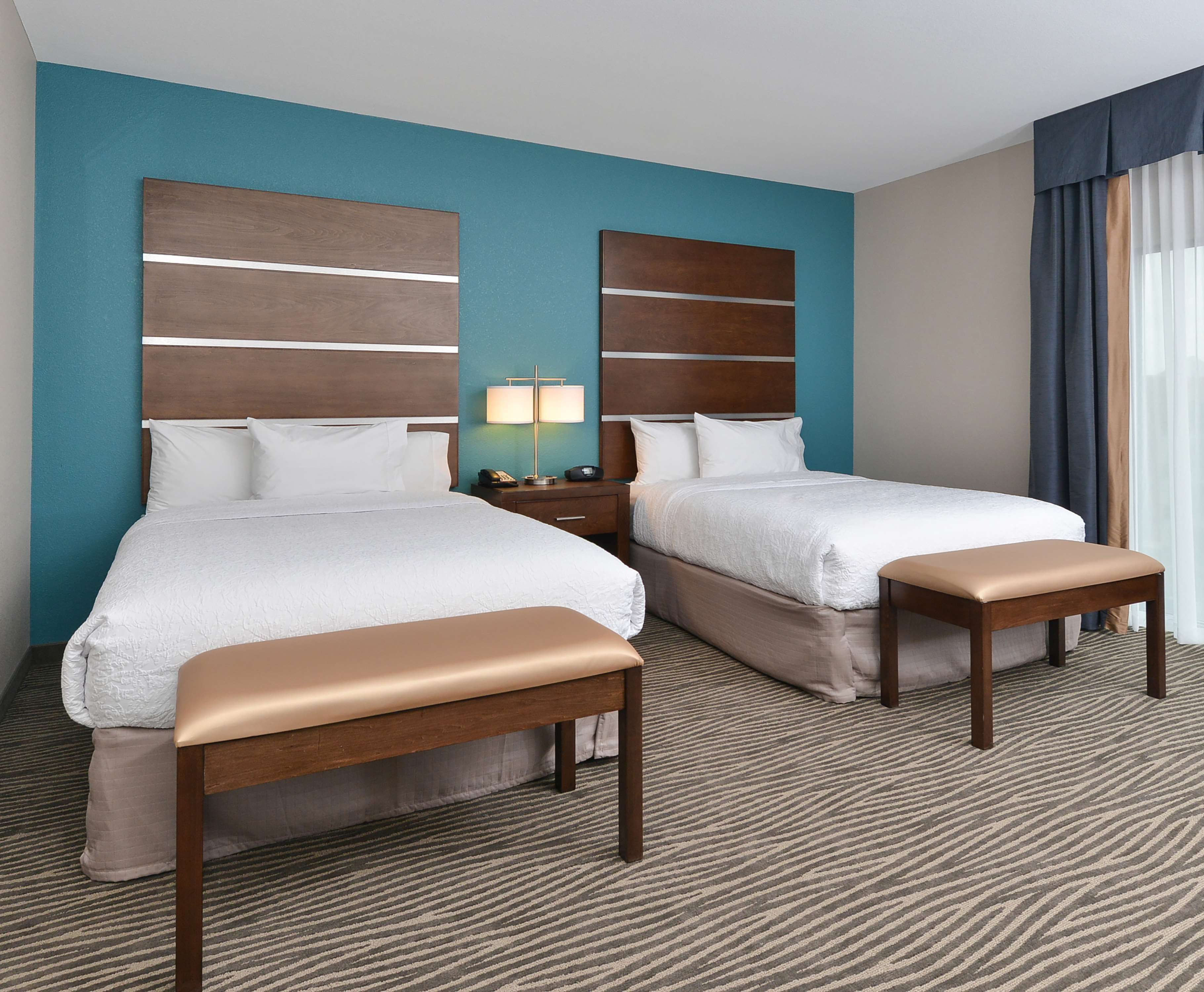 A hotel room with two beds, a bench, a nightstand with a lamp, and a view of the outdoors.