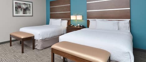 A hotel room with two beds, a bench, a nightstand with a lamp, and a framed picture on the wall.