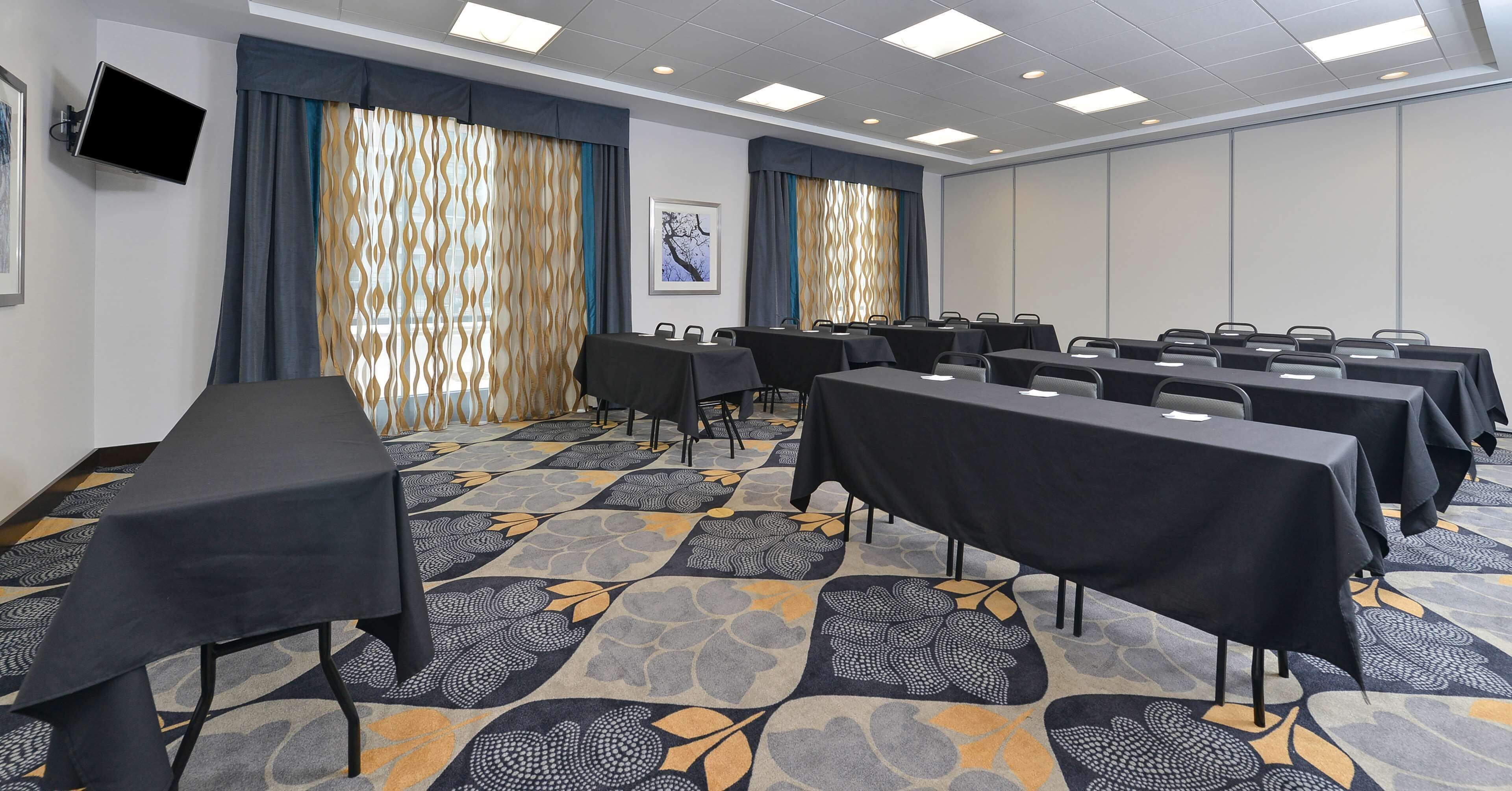 A conference room with multiple tables and chairs, patterned carpet, and a flat-screen TV mounted on the wall.