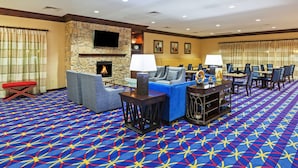 A hotel lobby with a stone fireplace, comfortable seating, a patterned carpet, and a dining area with tables and chairs.